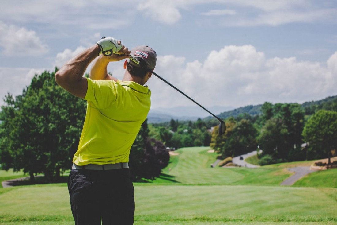 A golfer viewed from behind at the top of a swing, looking out over rolling countryside.